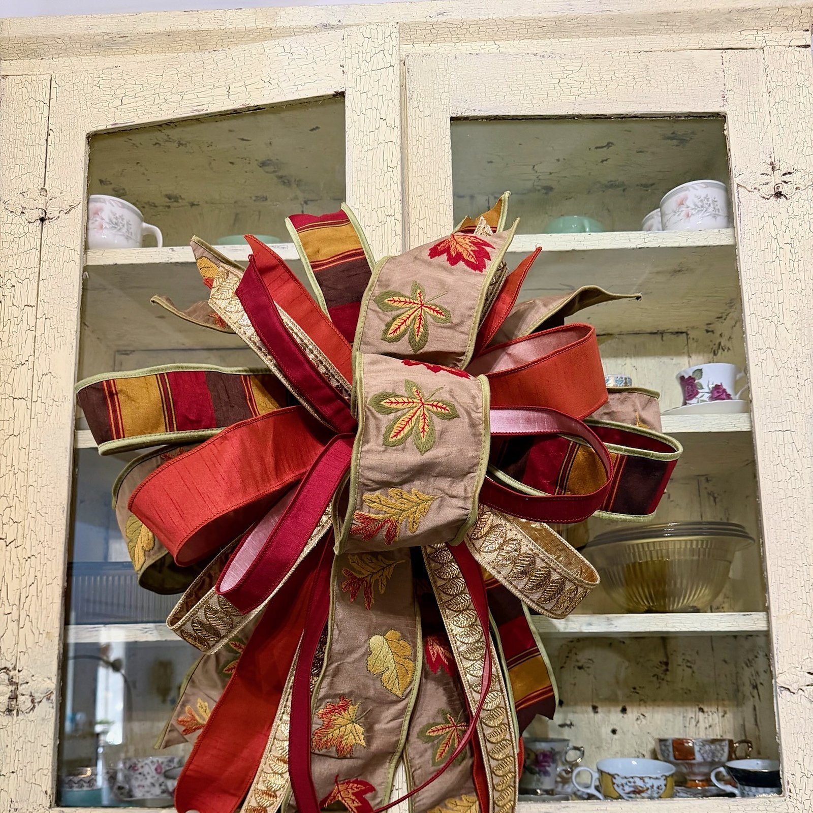Handmade bow in burgundy, rust, and gold striped ribbon with gold metallic leaf embroidery, hanging against a cream crackle-painted china cabinet